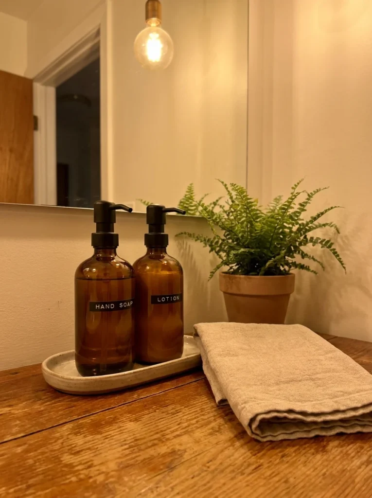 Matching glass pump dispensers for soap and lotion on a rental bathroom counter, warm bathroom lighting, vertical Pinterest photography, warm decanted styling